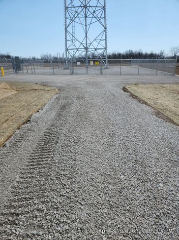 Gravel path leading to a fenced communication tower on a clear day.