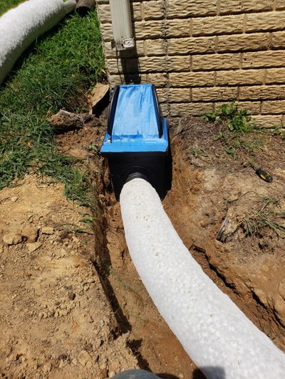 White drainage pipe connected to a blue and black drainage box near a brick wall.