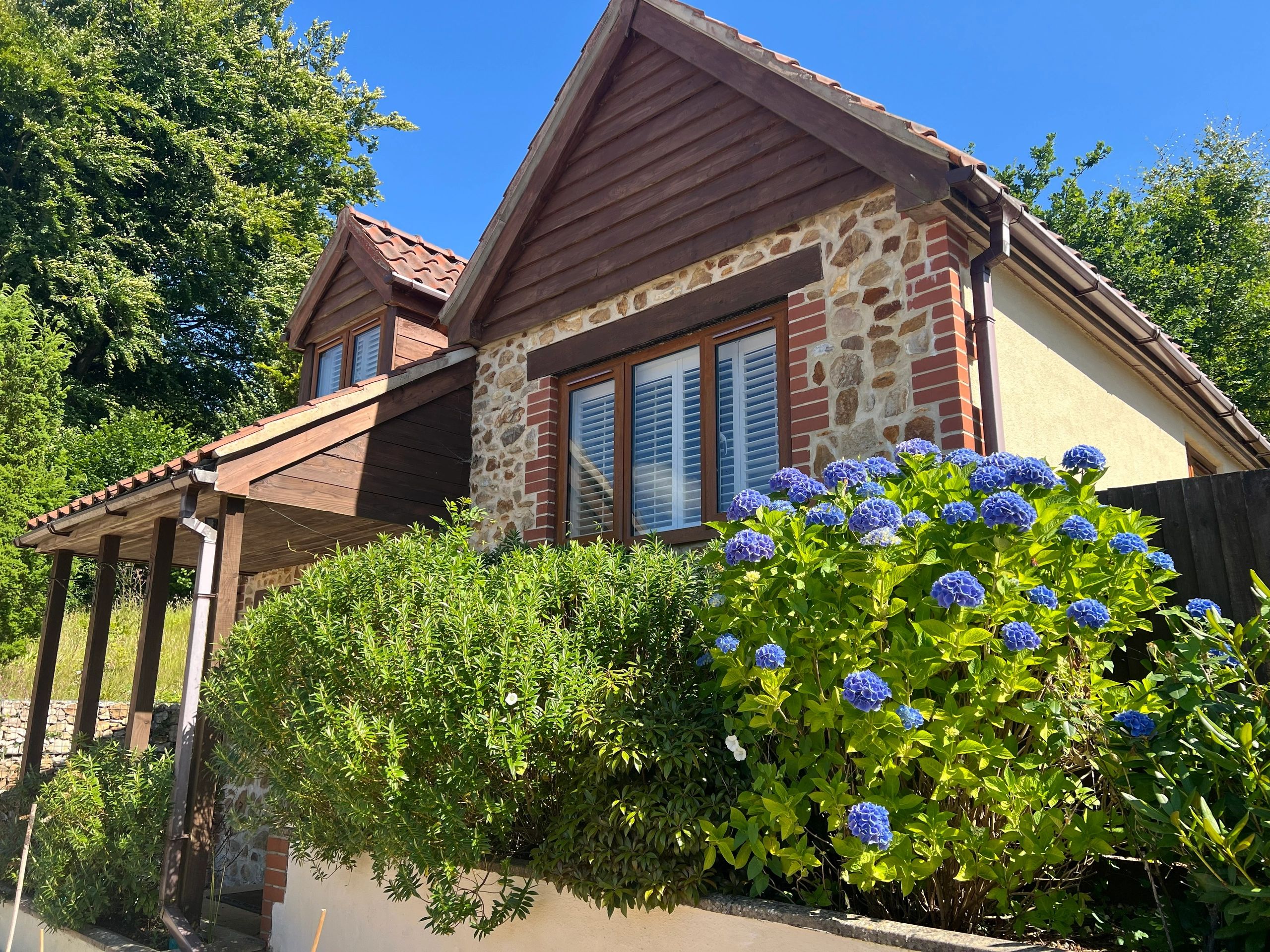 Cozy stone house with blue hydrangeas and greenery under clear blue sky.