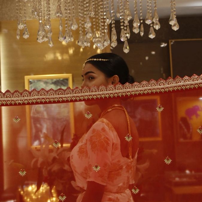 Woman in traditional attire behind a sheer red dupatta with hanging crystals above.