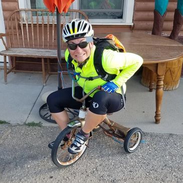 Adult woman on a small kids tricycle, wearing a bright yellow shirt & biking helmet.