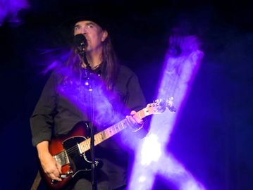 Musician playing electric guitar on stage with purple lighting.