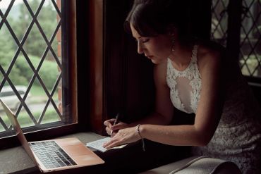 Woman writing notes near a window with a laptop.