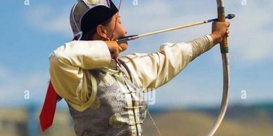 Person in traditional attire aiming a bow and arrow outdoors.