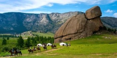 Large rock formation resembling a turtle in a green valley with horses and tents.
