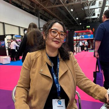 Woman with glasses and brown jacket sitting at an event with a pink carpet.