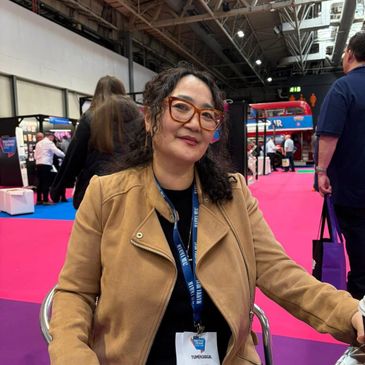 Woman with glasses and brown jacket sitting at an event with a pink carpet.
