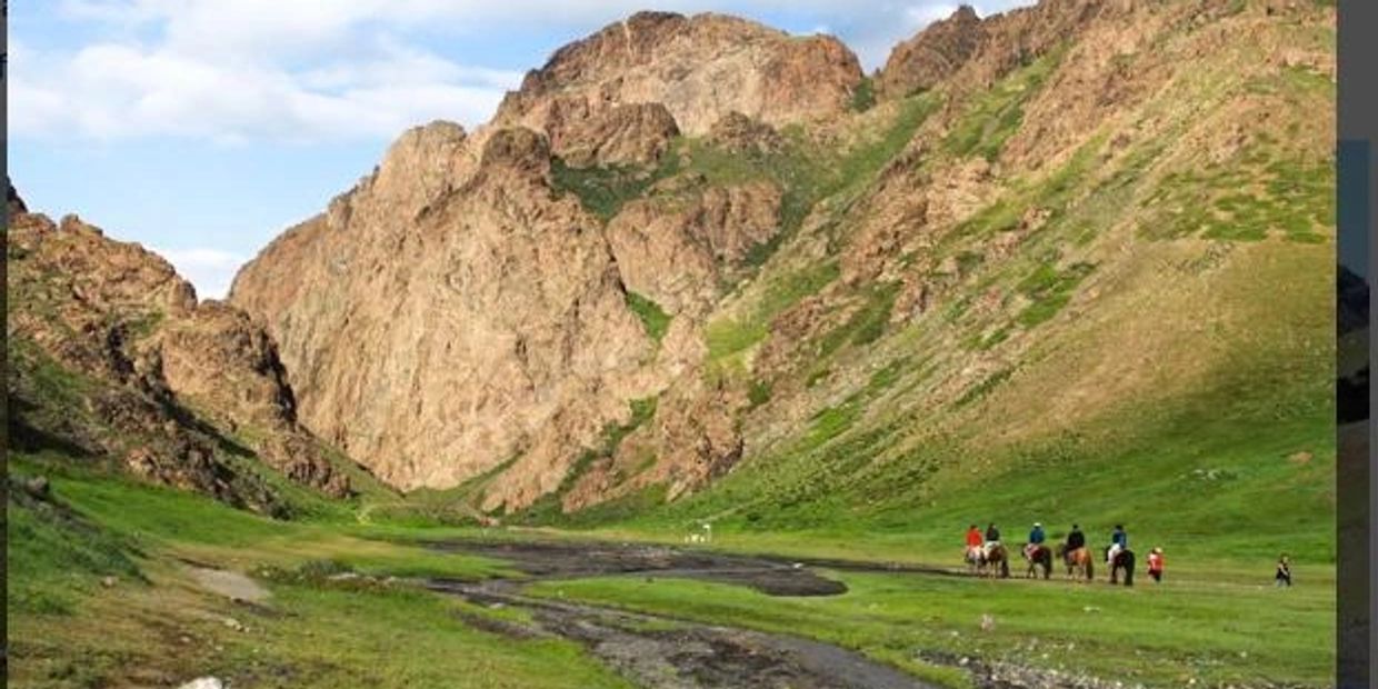 Group riding horses through a green valley with rocky hills.