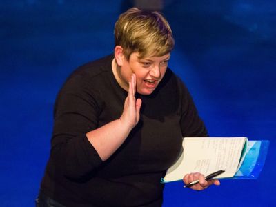 Photo of Sue Moore, in black with blue background, raising her hand to her face holding a notebook