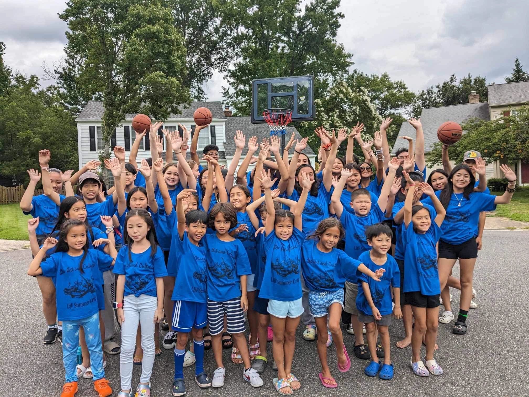Group of children in blue shirts raising hands outdoors near basketball hoop.