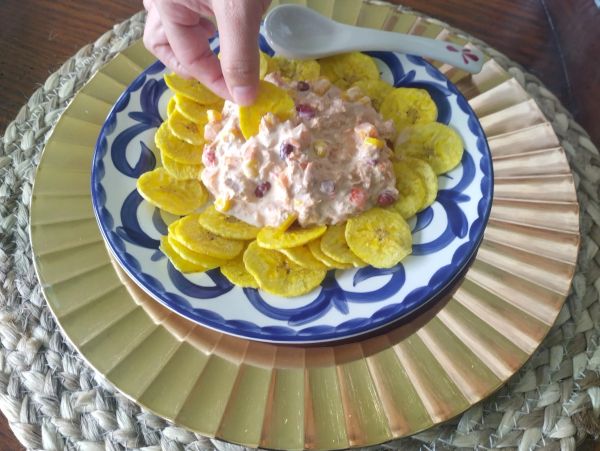 Hand dipping plantain chip into creamy dip on decorative plate.