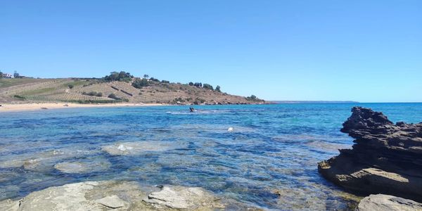 Clear blue sea with rocky shore and distant hills under a bright sky.