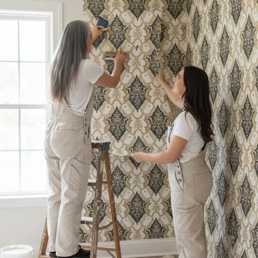 Two women in overalls hanging patterned wallpaper in a bright room.