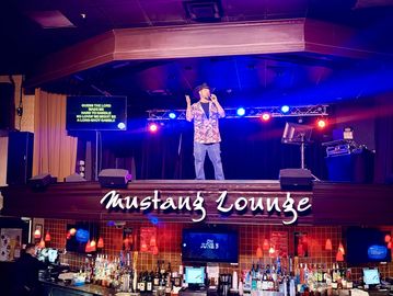 A man performing on stage at Mustang Lounge with karaoke lyrics displayed.