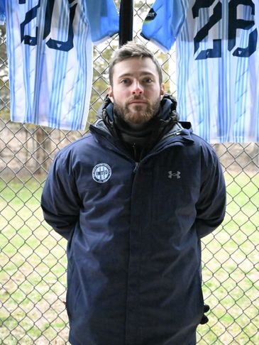 Man in a dark jacket standing in front of a fence with sports jerseys.