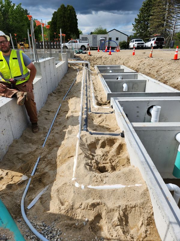 Construction site with concrete foundations and piping in sandy trench, worker in safety gear sitting nearby.