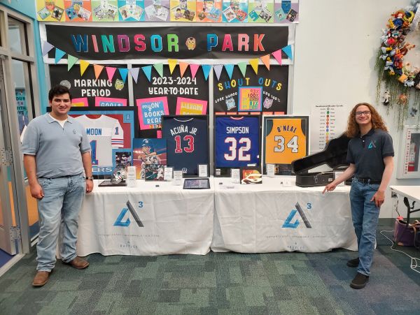 Two young men standing beside a display table with sports memorabilia and Windsor Park banners.