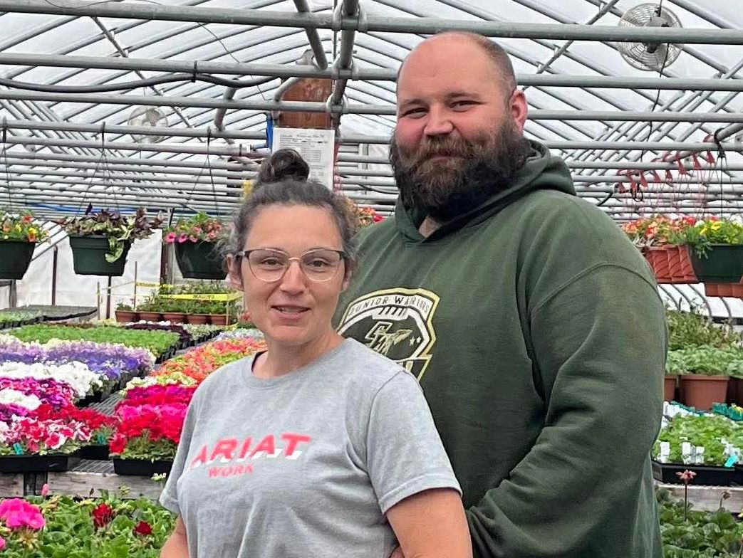 A smiling couple stands inside a greenhouse surrounded by colorful flowers and plants.