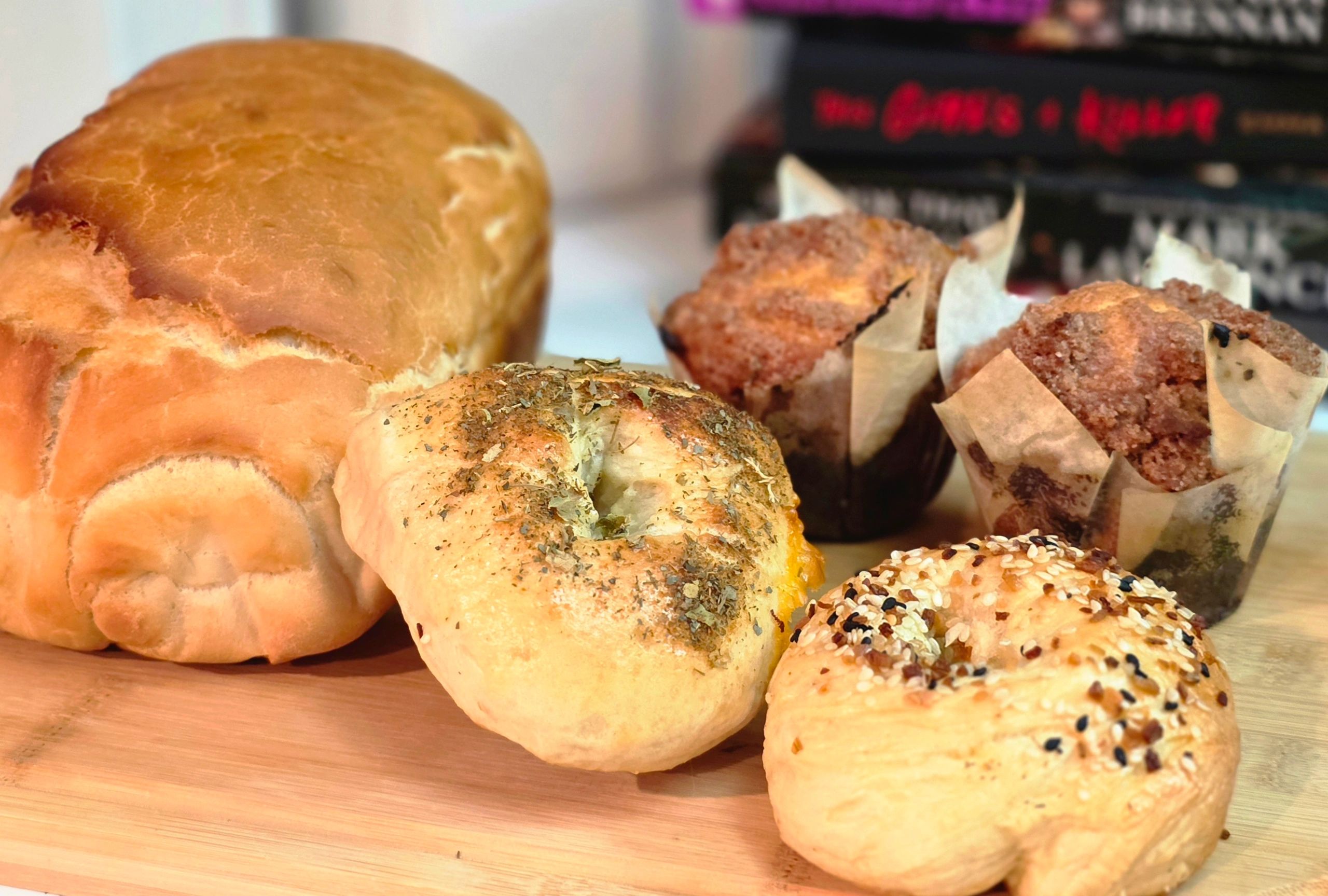 A loaf of freshly bakes bread, bagels and blueberry muffins sitting on a cutting board.
