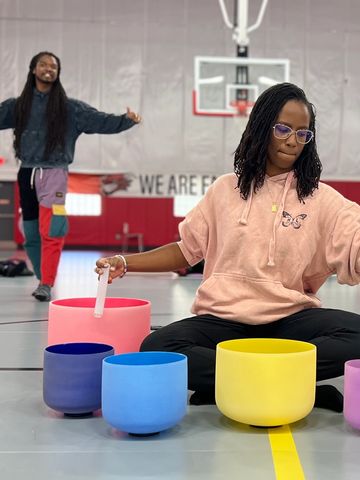 Woman playing colorful singing bowls in a gymnasium while a man dances in the background.