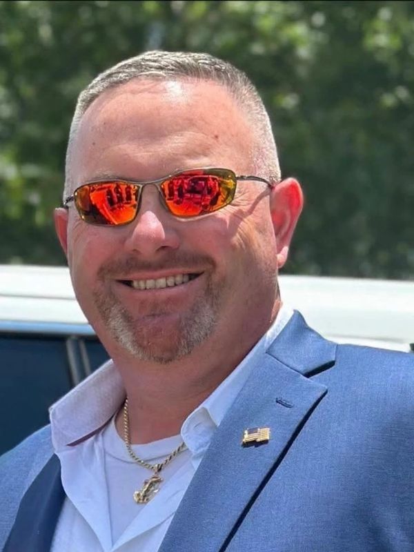 Smiling man in a blue blazer and reflective sunglasses outdoors.