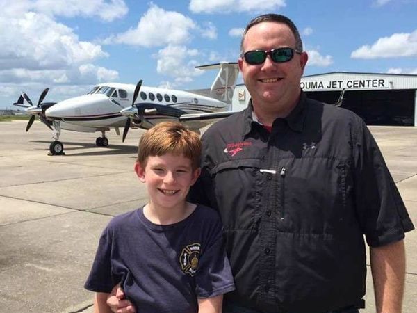 Father and son smiling together at an airport with a small plane behind them.