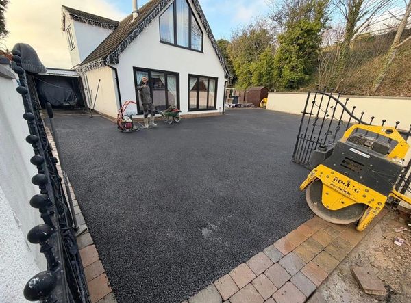 Newly laid black asphalt driveway in front of a modern house with a worker standing nearby.