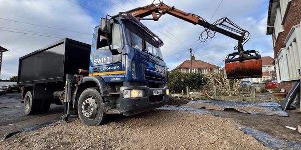 Blue Swift truck with an excavator arm unloading gravel near houses.