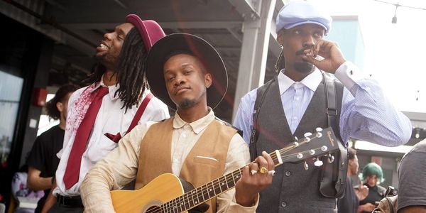 Three stylish men, one playing guitar, enjoying a lively outdoor moment.