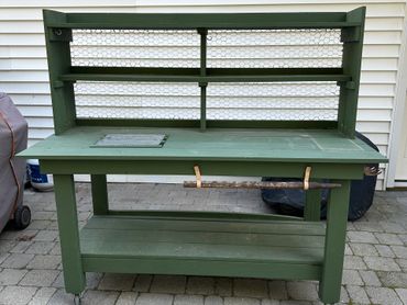 Green wooden potting bench with shelves and a stick on a patio.