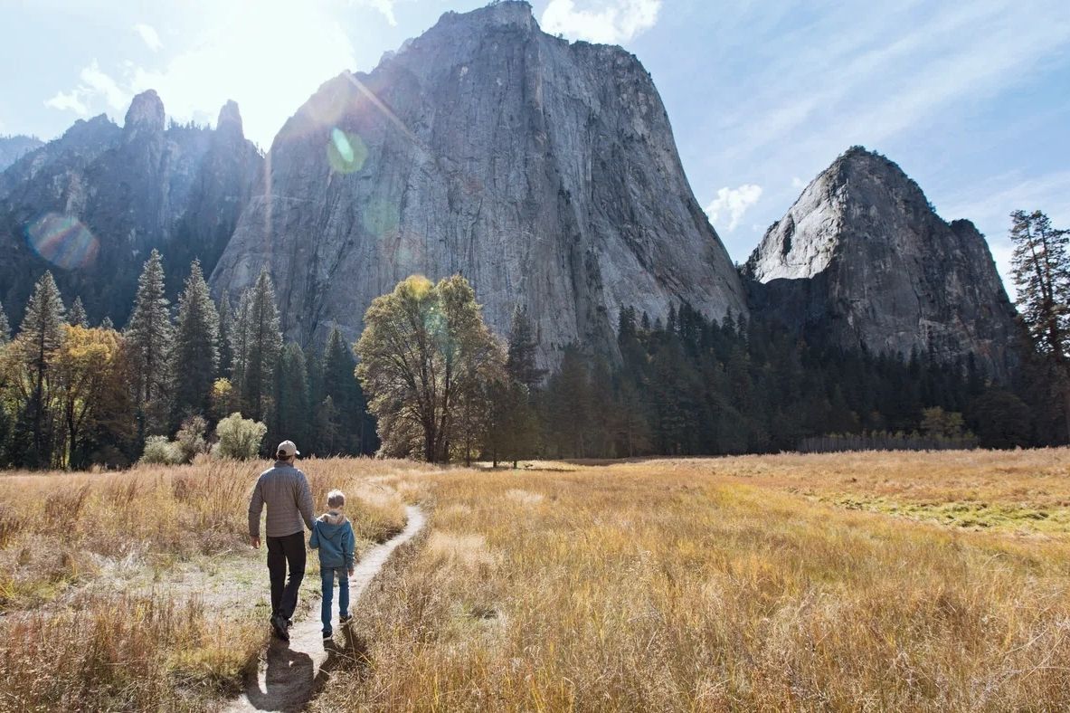 Image Description: A parent and child walking on a dirt path through a grassy field towards tall mou