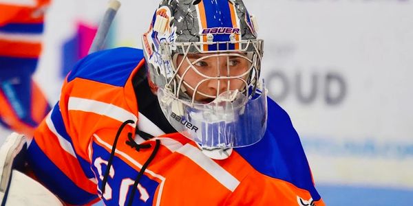 Hockey goalie wearing orange and blue gear prepping for a save.