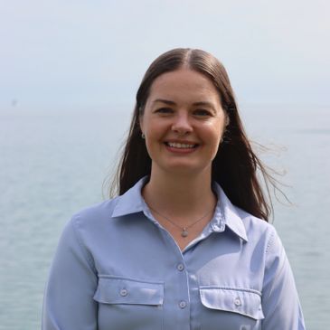 Smiling woman with long hair standing by the water in a light blue shirt.