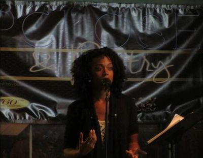 A woman speaks into a microphone on stage with a dark backdrop.