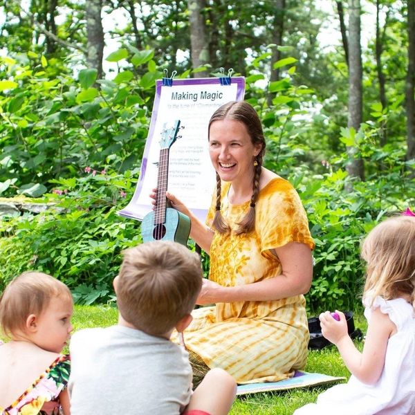 A woman in a yellow dress teaches children outdoors with a ukulele.