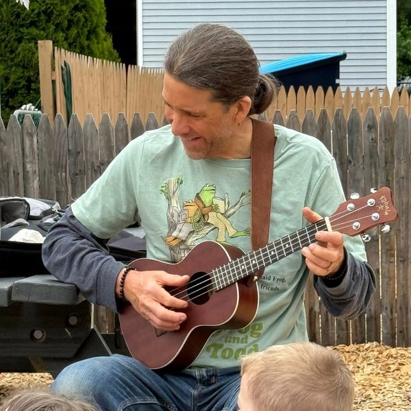 Man playing a ukulele outdoors with children nearby.