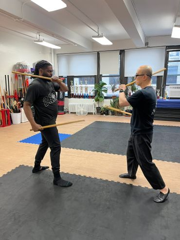 Two men practicing martial arts with wooden sticks in a bright training room.