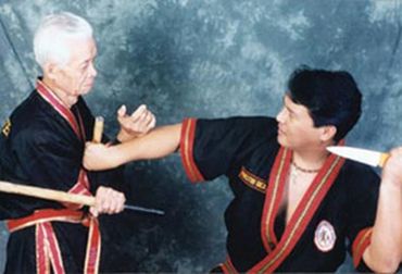 Two martial artists in black uniforms practicing self-defense against knife attacks.