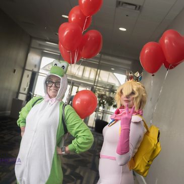 Two cosplayers dressed as Yoshi and Princess Peach holding red balloons indoors.