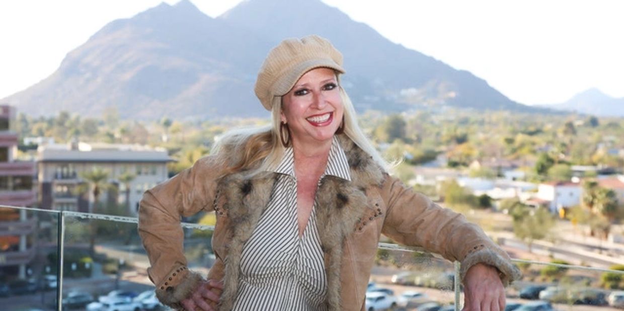 Smiling woman in a fur-lined jacket and hat poses outdoors with mountains in the background.