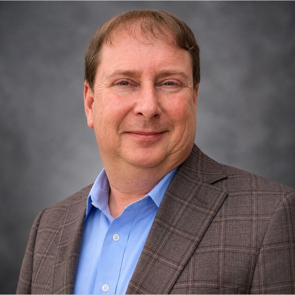 Man in a brown plaid blazer and blue shirt smiling.