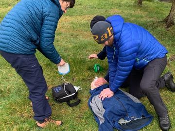 2 first aiders giving CPR. One is doing chest compressions whilst the other is preparing AED pads.