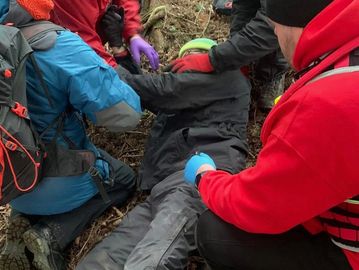 First aiders helping a casualty roll on to their side. The casualty is lying on the forest floor.