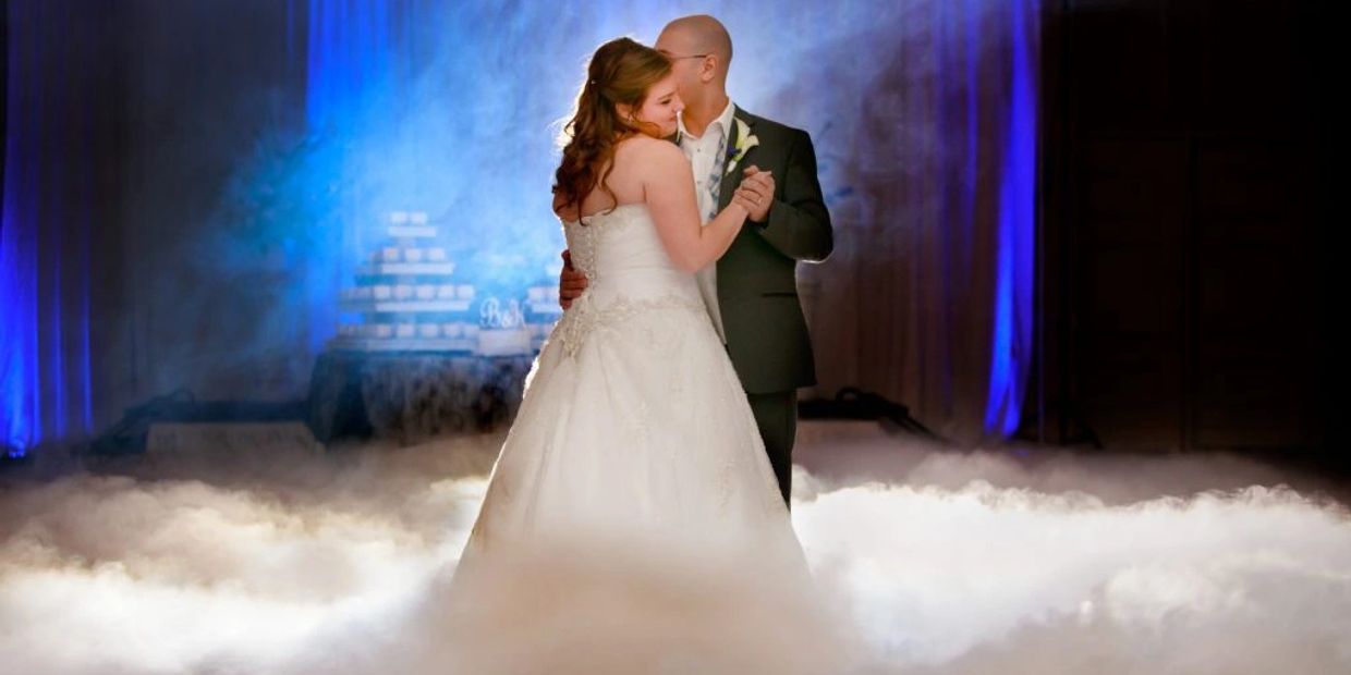 A bride and groom share a romantic first dance surrounded by fog and blue lighting.