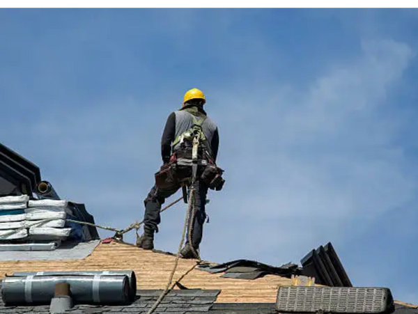 A roofer wearing safety gear works on a roof under clear sky.