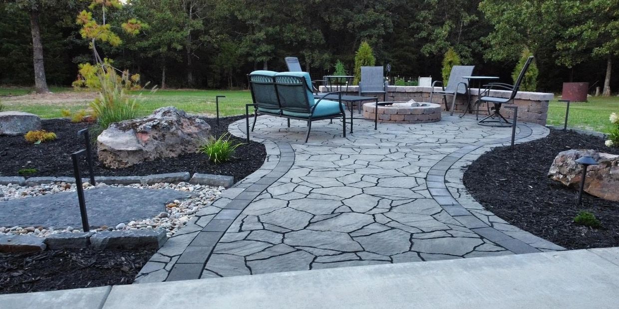 Outdoor paver patio with furniture, a firepit, landscaping garden, and a wooded area in background