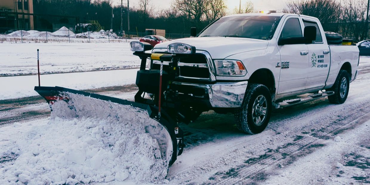 White truck with logo on the side and a plow in the front pushing snow in a commercial parking lot. 