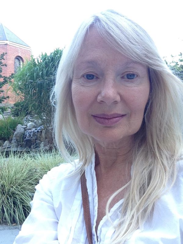 A woman with long blonde hair smiling outdoors near greenery and a brick building.