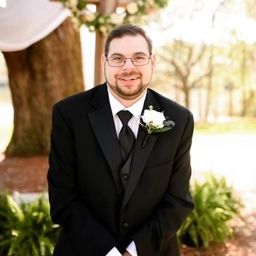 Smiling groom in black tuxedo with white boutonniere outdoors.