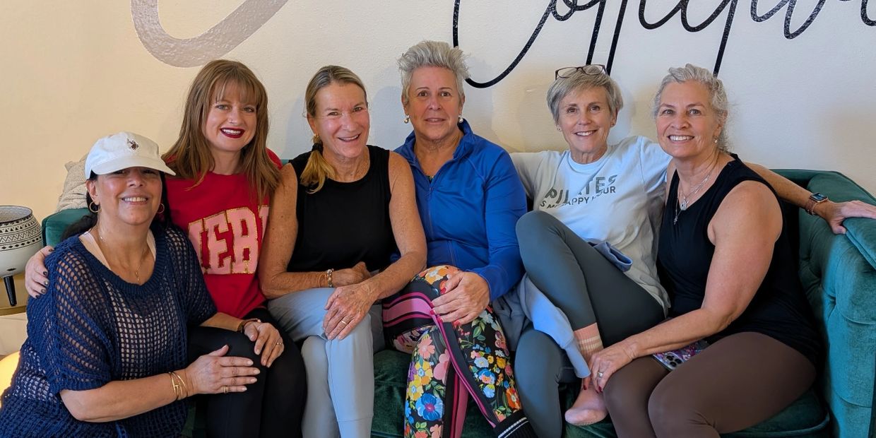 Six women sitting closely on a couch, smiling happily at the camera.